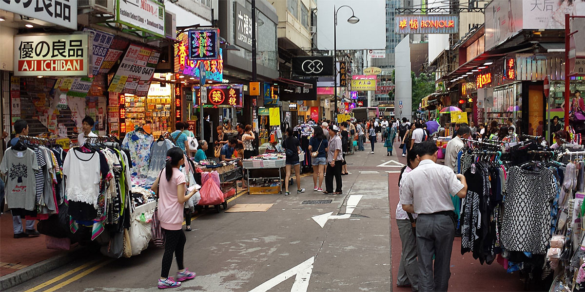 Bustling pedestrian street in Sneakers Street, Hong Kong, with clothing stalls, shoppers, and neon shop signs